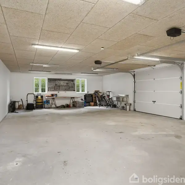 Garage med åben port indeholder værktøjsbænk, cykler og diverse redskaber. Loftslamper oplyser rummet, og et vindue i baggrunden giver naturligt lys. Betongulv og hvidmalede vægge fuldender rummet.