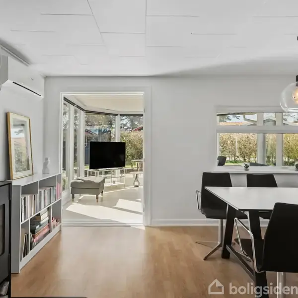 Brændeovn opvarmer stue med spisebord og stole. Store vinduer og åbning mod udestue med udsigt til have. Hængelamper lyser op ad rum med reoler og billeder.