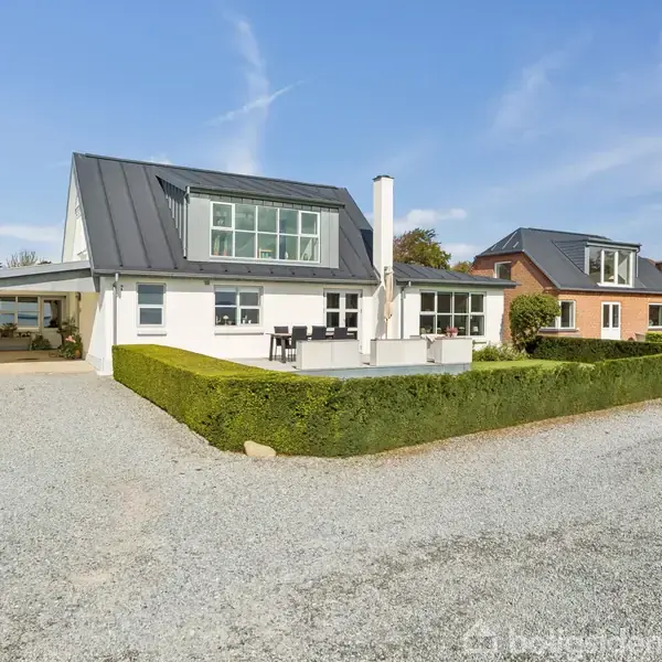Hvidt hus med sorte tagvinduer, omkranset af hæk og grussti. Foran huset er der en terrasse og grusbelagt indkørsel. Blå himmel og anden bolig ses i baggrunden.