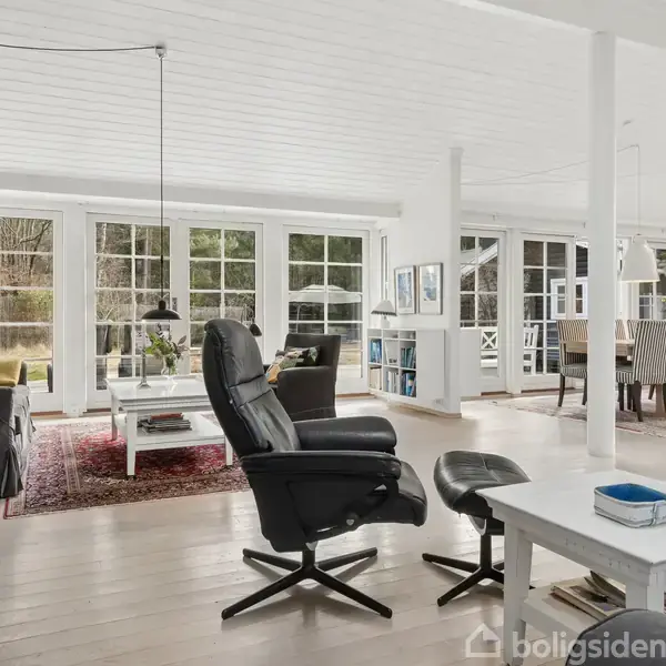 Stue med sort lænestol ved siden af et sofabord; ild brænder i pejsen. Store vinduer bringer lys ind, og spiseplads med langbord ses i baggrunden.