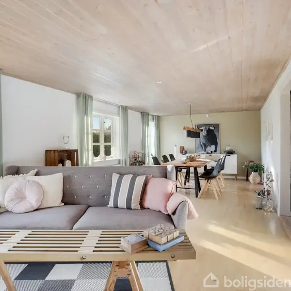 Grå sofa med puder står i en lys stue. Foran sofaen er et sofabord med bøger og dekorationer. Rummet har lyse trælofter og stort spisebord i baggrunden.