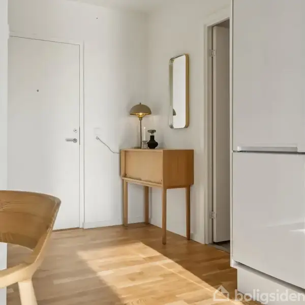 En træstol står ved et rundt glasbord; en kommode med lampe og spejl står langs væggen i et lyst rum med parketgulv og en lukket dør.