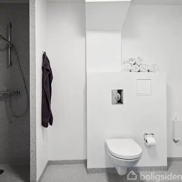 Bruser og håndklæde hænger til venstre, væghængt toilet med toiletpapirholder og rullede håndklæder på hylde til højre. Minimalistisk badeværelse i lysegrå og hvide farver.