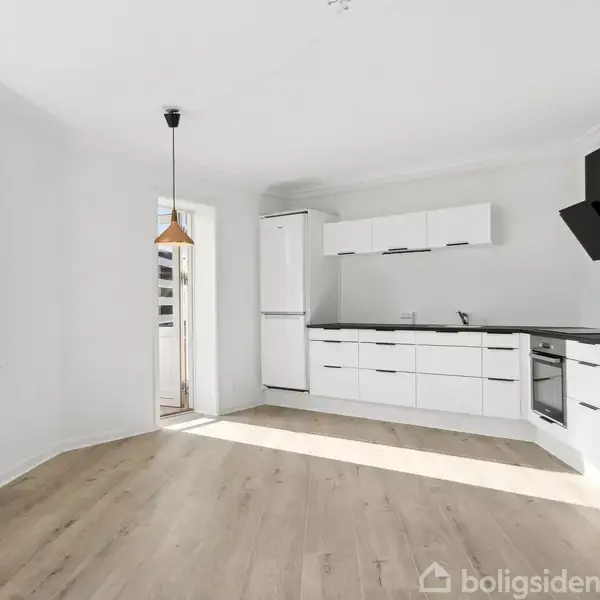 Et moderne køkken med hvide skabe og sorte bordplader i aktion, placeret i en solrig, hvidmalet stue med trægulve. En dør åbner til en lille altan.