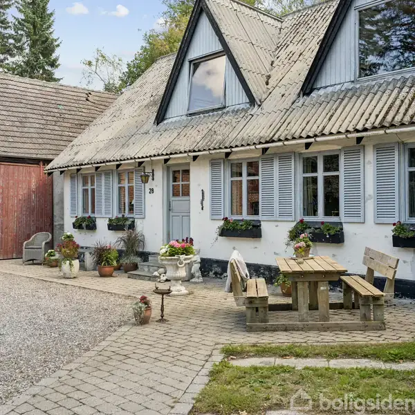 Hus med hvid facade og blå skodder, blomsterkasser under vinduerne. Et lille træbord med bænke står på terrassen. Dør markeret med tallet "28". Gruset gårdsplads omgiver huset.