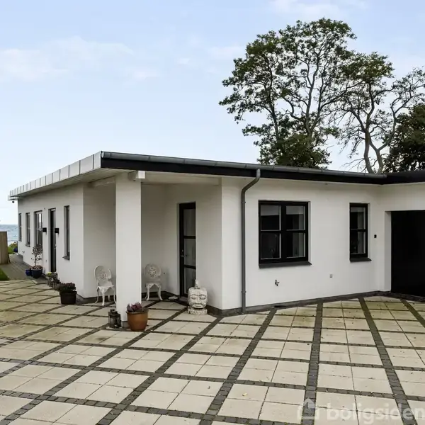 Hvid, moderne étplansvilla med fladt tag står ved flisebelagt indkørsel. Plantekrukker pryder terrassen, mens store træer og havudsigt skaber en naturskøn baggrund.
