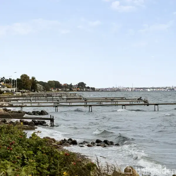Strand med bølger, moler strækker sig ud over vandet. Kystlinjen er beplantet med græs og buske, mens huse kan ses i baggrunden langs vejen.
