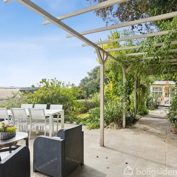 En terrasse med moderne havemøbler står under en pergola dækket af grønne planter. Til venstre ses et spisebord, mens marker strækker sig i baggrunden under en klar himmel.