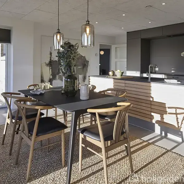 Spisebord med træstole står på et tæppe i en moderne spisestue. Pendellamper hænger over bordet. Køkkenet i baggrunden har sorte skabe og en lys træfrontet bar.