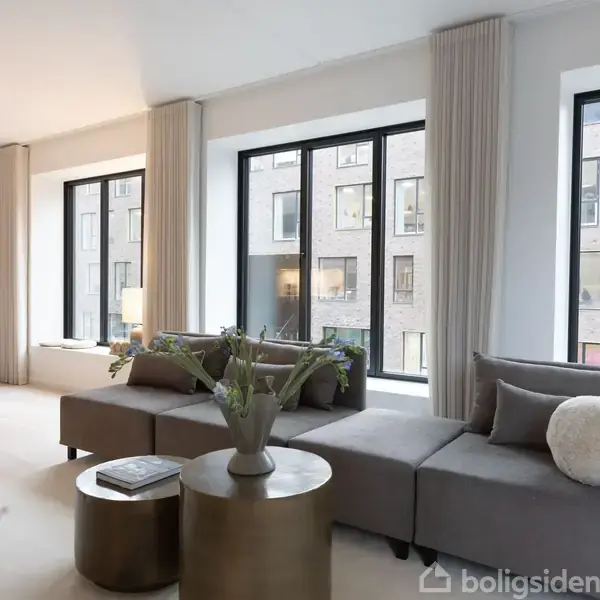 En grå sofa står i en lys stue, dekoreret med puder og planter. Bagved er store vinduer med udsigt til en murstensbygning. Der er gulv til loft gardiner ved vinduerne.