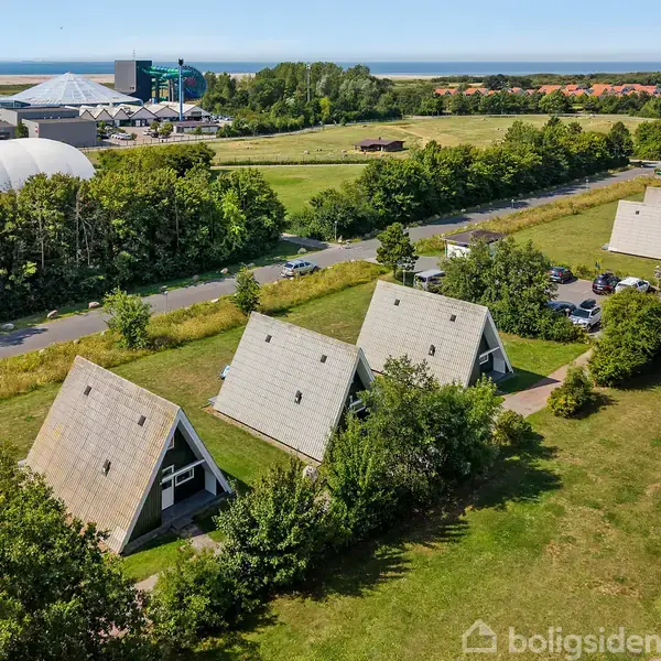 Flere A-formede sommerhuse står på en grøn mark. Vejen med parkerede biler løber mellem husene og træerne, mens afstand til havet ses i baggrunden under en klar himmel.