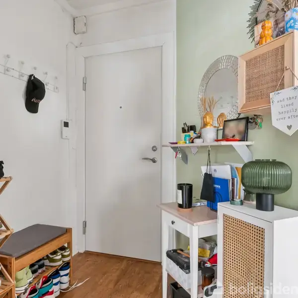 Dør i en entré med bænksæt, skohylde fyldt med sko. Hylde på væg med dekorationer og en seddel: “THE HAPPY LOVE FAMILY FINDS A WAY.” Væg malet lys grøn.