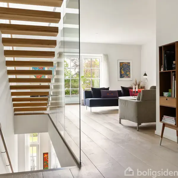 Trappen i træ svæver op ad væggen ved siden af en glasvæg. I baggrunden findes en stue med sofaer, et bord og et skab i et lyst rum med store vinduer.