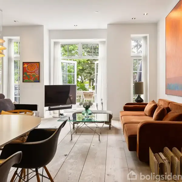 Stue med brun sofa og tv, opstillet ved et glasbord. Store vinduer og åbne døre til en have. Moderne kunst på væggene og spisebord med ikonisk lys.