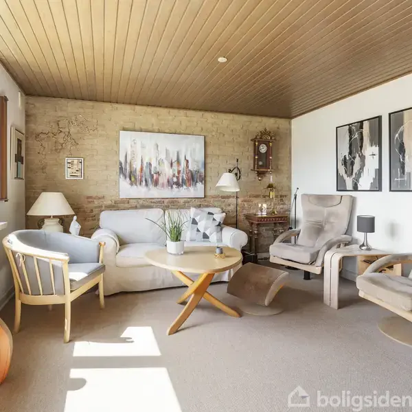 Stue møbleret med en beige sofa, to lænestole, og et sofabord. Væggen har moderne kunst. Der er en stor brikvæg, træloft, klokke på væggen, og vinduer med dagslys.