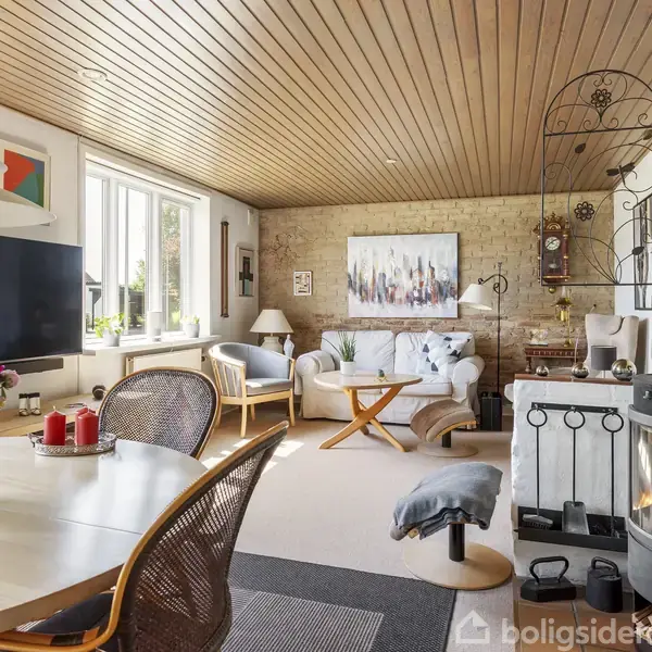 Stue med rund spisebord (med blomster og lys) til venstre. Sofa og lænestol står på et tæppe ved et sofabord. Brændeovn til højre skaber hygge. Lysindfald fra vinduerne.
