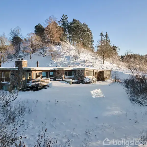 Et moderne hus står dækket af sne. Huset er omringet af et bakket landskab med træer, og vejret er klart med blå himmel.