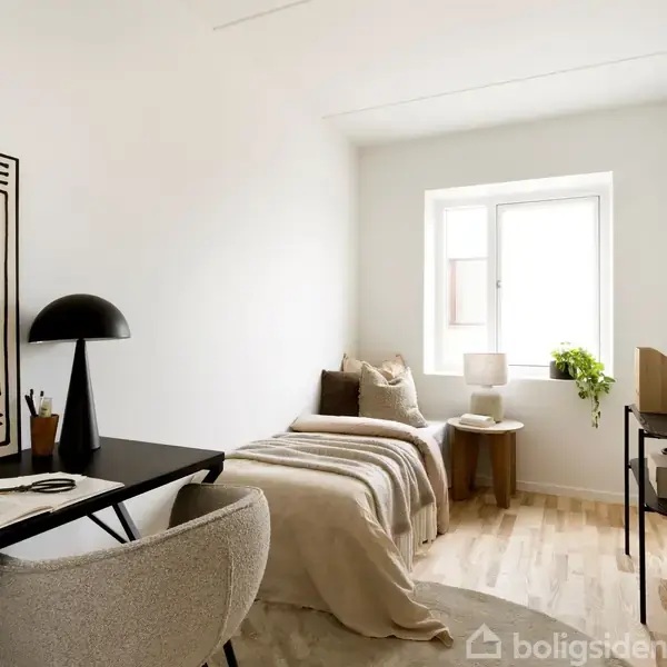 Et lille værelse med enkeltseng dækket med beige sengetæppe ved vindue. Ved siden af er et natbord med lampe og plante. Skrivebord modsat, med stol og lampe.
