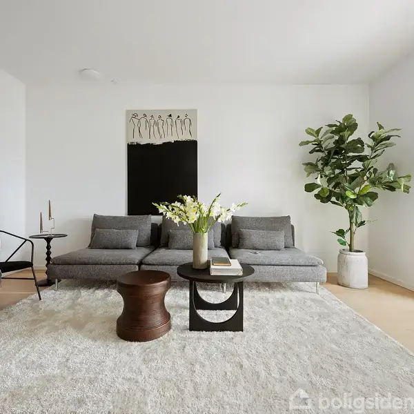 En grå sofa med puder står i et moderne, lyst stueområde. På gulvet ligger et blødt tæppe, omgivet af et sidebord og en gulvplante. Kunst hænger på væggen.