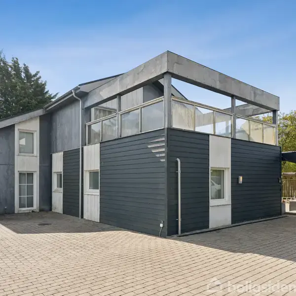 En moderne bygning står stille, omgivet af en flisebelagt gårdsplads og frodig vegetation i baggrunden. Bygningen har en grå og hvid facade med store vinduer og en carport til højre.