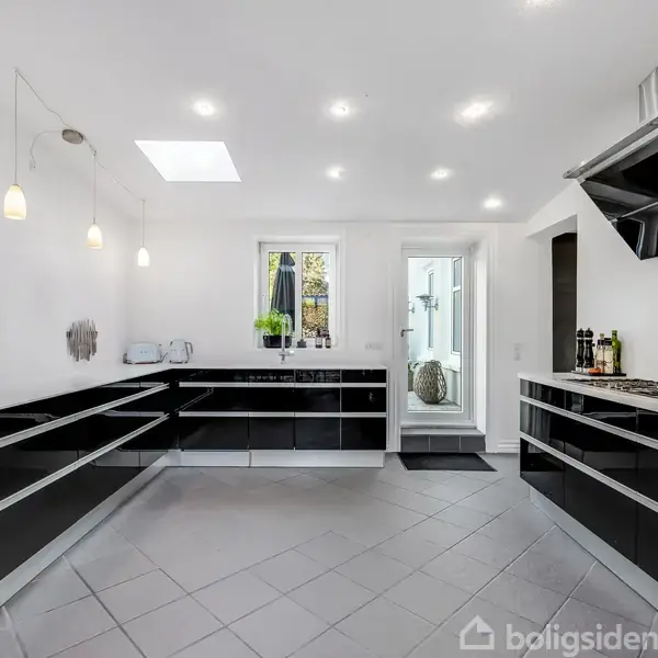 Et moderne køkken med sorte skabe og stålgreb står tomt. Loftslamper og naturligt lys fra vinduet oplyser rummet, der har grå flisegulve. En dør fører til en anden del af huset.