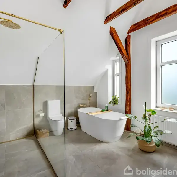 Brusekabine med glasvæg, gulv i lyse fliser, ved siden af et fritstående karbad og toilet. Vindue på højre væg, store bjælker i loftet, grønne planter ved siden af.