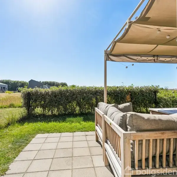 Træmøbler står på en flisebelagt terrasse under en pergola, omkranset af en lav hæk. I baggrunden ses åbne marker og et hegn, med flere bygninger i det fjerne.