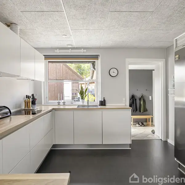 Et moderne køkken med hvide skabe og træbordplader. En brødrister og knivblok står på bordet. Et vindue ud til en gårdsplads. En dør fører til en gang med jakker.