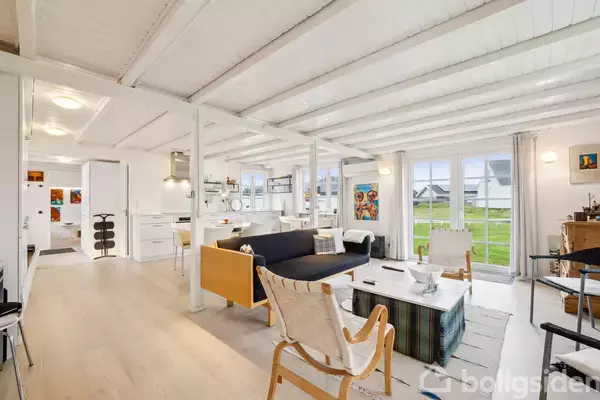 Stue med moderne møblering; sofa og stole omkring et sofabord. Køkken i baggrunden. Store vinduer giver udsigt til en grøn, græsbeklædt have. Loftsbjælker og lyse træelementer dominerer.