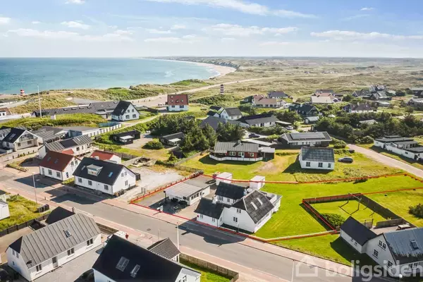 Huse står stille i en lille kystlandsby med sandklitter og havet i baggrunden. Husene er spredt langs en vej, mens en markeret grund er synlig midt i billedet.