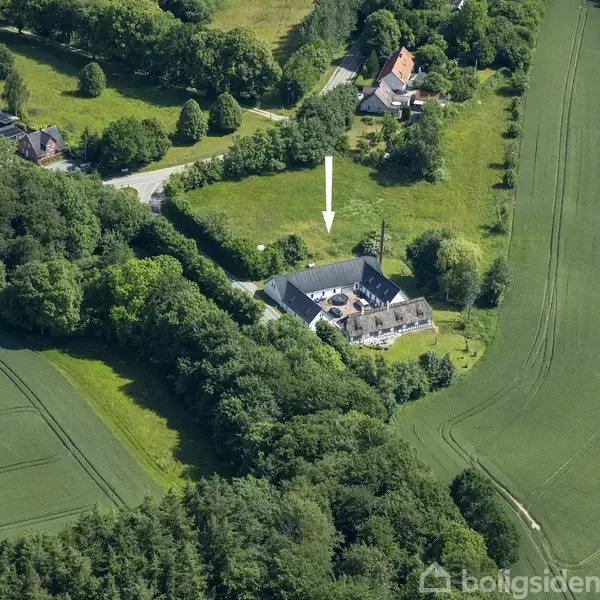 Et stort hus med en gårdsplads vises fra luften med en hvid pil pegende på det. Det er omgivet af marker, skov og enkelte andre huse.