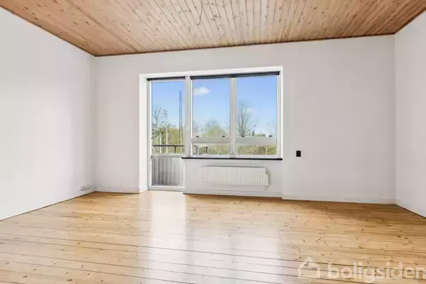 Et tomt rum med lyse trægulve og træbeklædt loft. Store vinduer og en dør åbner ud mod en balkon, hvor man kan se trætoppe udenfor. Ingen tekst i billedet.