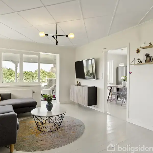 En moderne stue med en grå sofa som står i et hjørne og bøjer mod et sofabord. En stor fladskærm er monteret på væggen. Store vinduer lader naturligt lys ind.