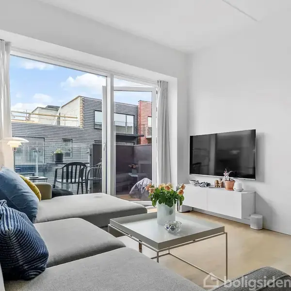 Lys stue med grå sofa foran stort vindue med udsigt til terrasse. Et sofabord med blomster står foran fjernsyn på væggen. Vægur hænger til højre.