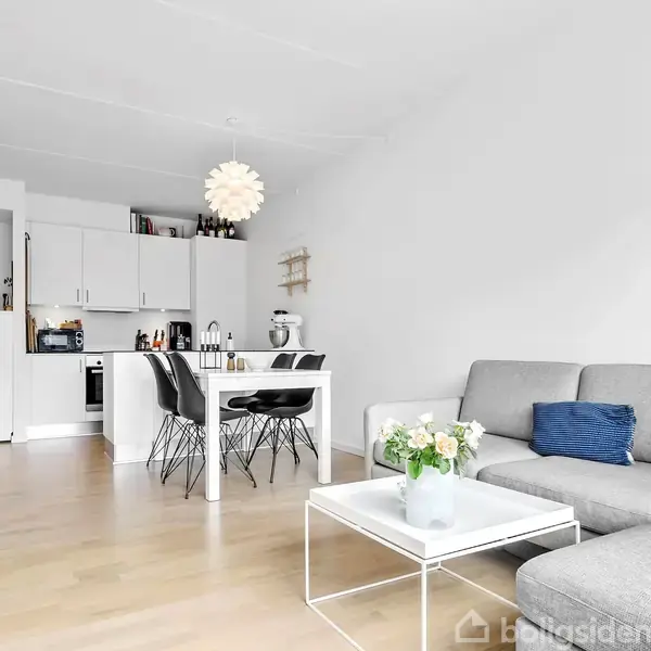 Stue med gråt sofaarrangement omkring et sofabord med blomster, ved siden af en spiseplads med hvidt bord og sorte stole; køkken i baggrunden med hvide skabe.