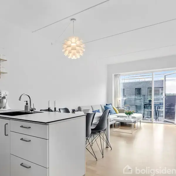 Køkkenområde med hvidt skab, vask og køkkenmaskiner. Ved siden af er en stue med sofa og sofabord. Store vinduer åbner mod en altan. Lampe hænger fra loftet.