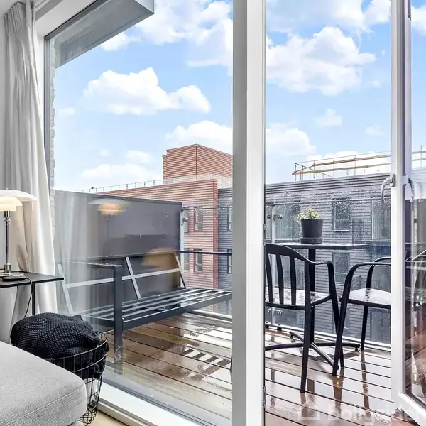 En sofa står ved en stor glasdør, der åbner ud til en balkon med to sorte stole og et lille bord. Bygninger og himmel ses i baggrunden.