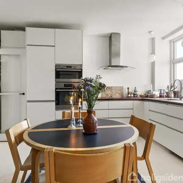 Rundt spisebord med træstole står midt i et lyst køkken. Bordet har en vase med blomster og tændte stearinlys. I baggrunden ses ovne, emhætte og store vinduer.