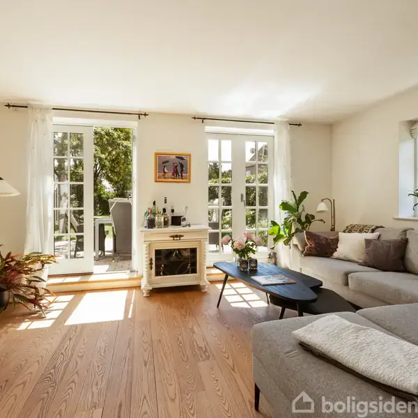 Stue med stor grå sofa, sofabord og fladskærms-tv. Sollys strømmer ind gennem franske døre, der fører til en terrasse. Planter og maleri skaber hyggelig atmosfære.