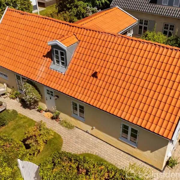 Et hus med orange tegltag ligger i en have med flisebelagt sti. Der er buske, små terrasser og et bord med stole omkring.
