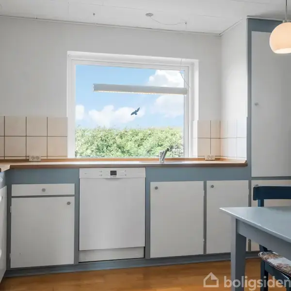 Køkken med hvide skabe og blålige kanter, set foran vindue med udsigt til grønne træer og blå himmel. Der er en ovn, opvaskemaskine og en spiseplads med bord og stol.