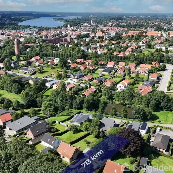 Landskab med huse i forgrunden omgivet af grønne træer, forbindelse til større by med tættere bebyggelse og en stor sø i baggrunden. Tekst: "773 kvm".