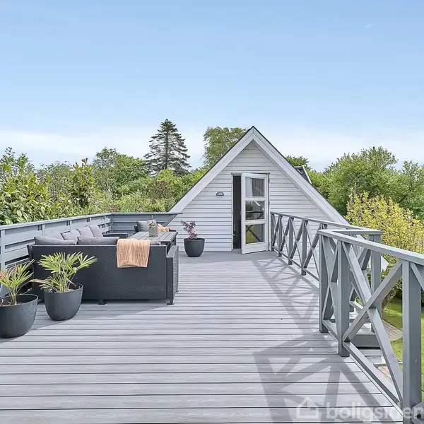 Et stort træterrasseområde med sofagruppe og blomsterkrukker. Terrassen er omgivet af et trægelænder og ligger højt over en frodig, grøn have.