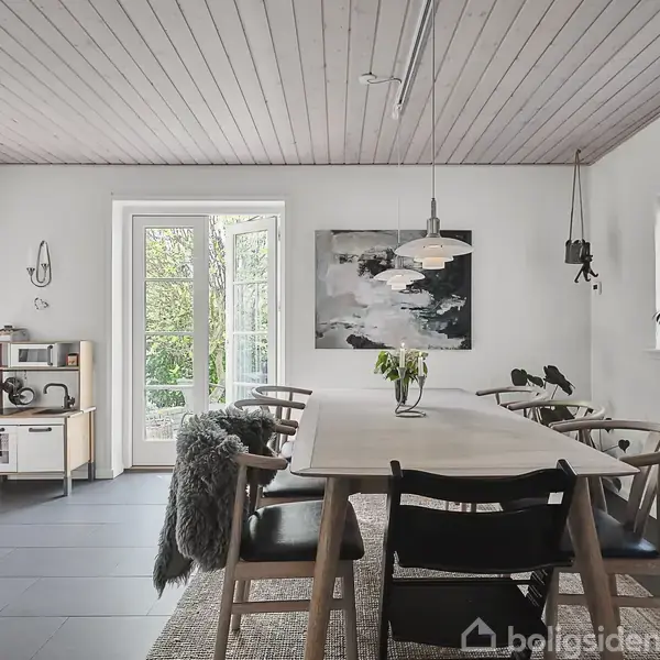 Et spisebord med stole er centralt placeret i en lys spisestue. Et maleri hænger på væggen, og store vinduer åbner ud til en frodig have. En lampe hænger over bordet. En lille reol står til venstre.