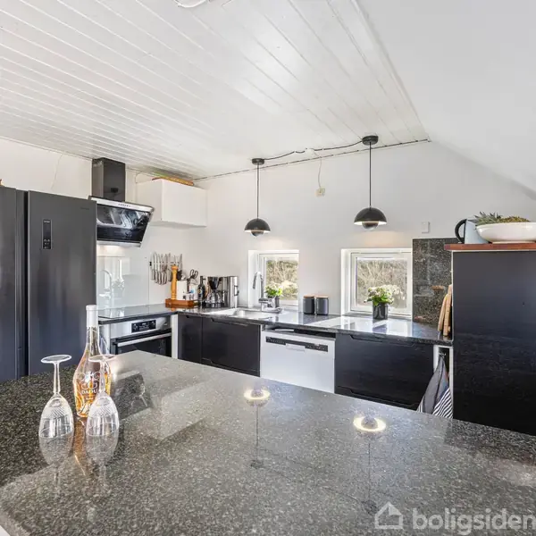 Køkken med sorte skabe og moderne apparater. Bordpladen er af mørk granit med lysestager. To små vinduer og loftlamper giver lys, mens blomster pynter rundt omkring.