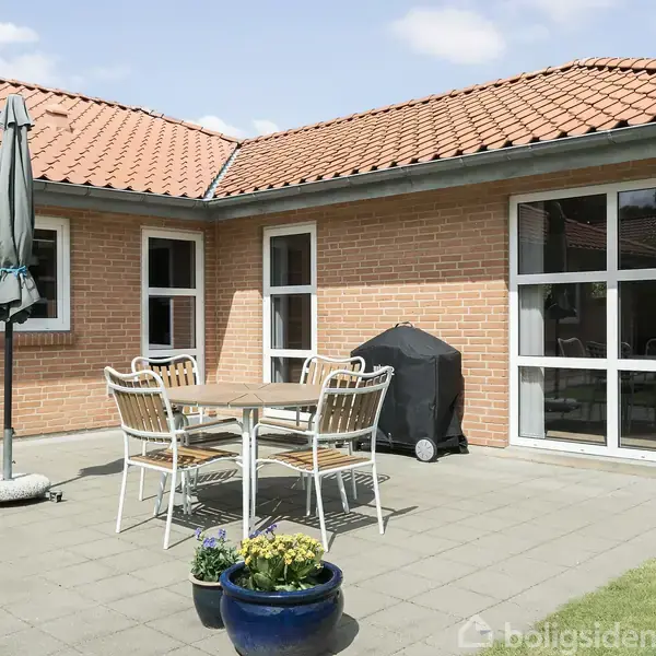 Et havebord med stabelstole omkring står på en terrasse ved et murstenshus. Parasol og grill ses i baggrunden, omgivet af blå blomsterpotte og græsplæne.
