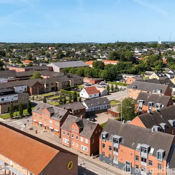 Et boligområde med røde murstensbygninger og orangerøde tage ligger stille. Bygningerne er tæt placeret langs gaderne, omgivet af grønne træer. I baggrunden ses flere huse og vegetation.