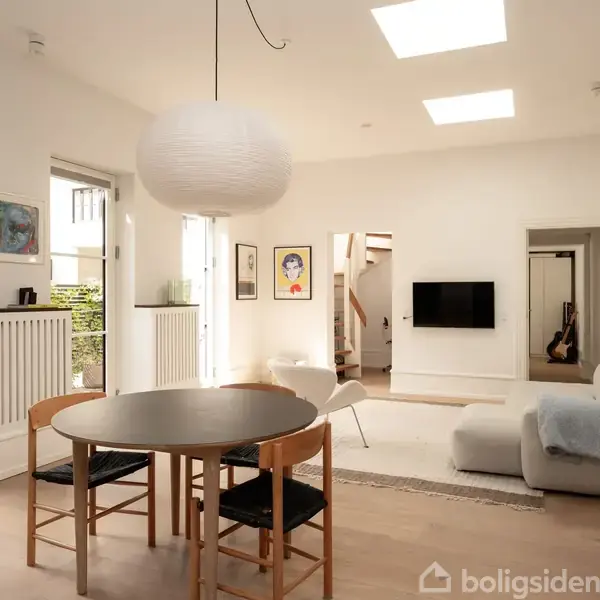 Et lyst køkken-alrum med spisebord og stole i forgrunden, en sofa og tv på væggen i baggrunden. Rummet er minimalistisk indrettet med kunst på væggene og hylder til højre.