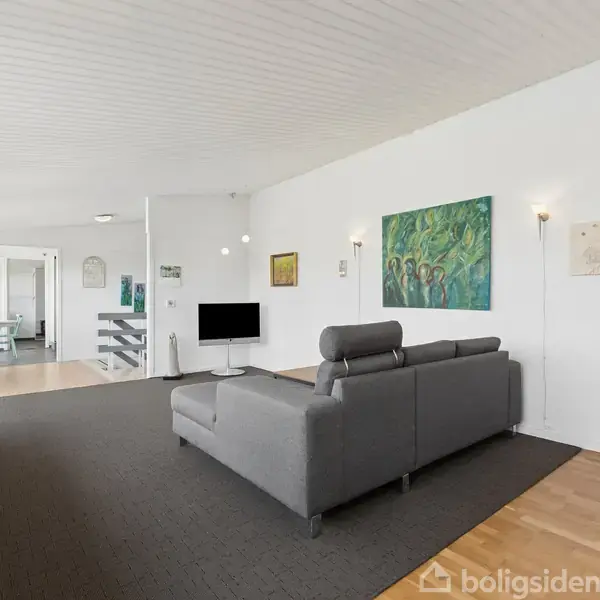 Grå sofa står på mørkt tæppe i en lys stue med trægulv. Væggen har farverige malerier, og et tv er monteret. Store vinduer giver udsigt til det udendørs, mens trappe leder til køkken.