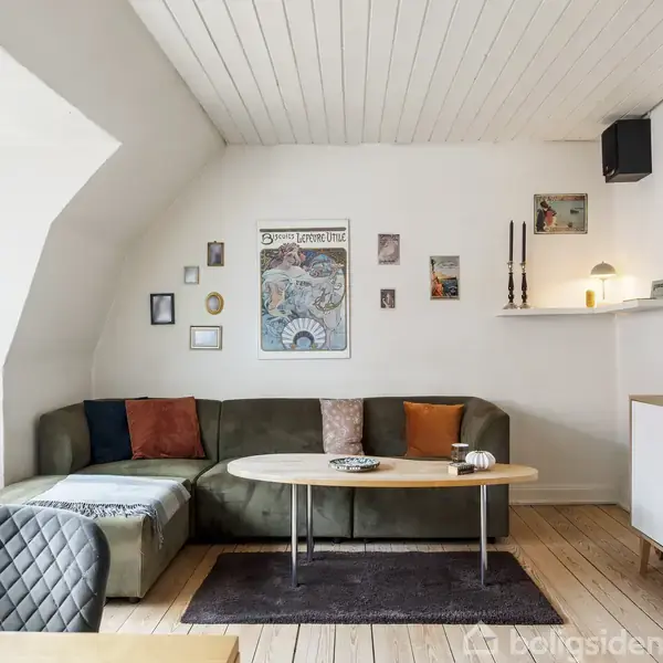 En stue med en grøn vinkelsofa fyldt med puder, står omkring et ovalt træbord. Væggene er udsmykket med kunst og hylder. Gulvet er af lyst træ.